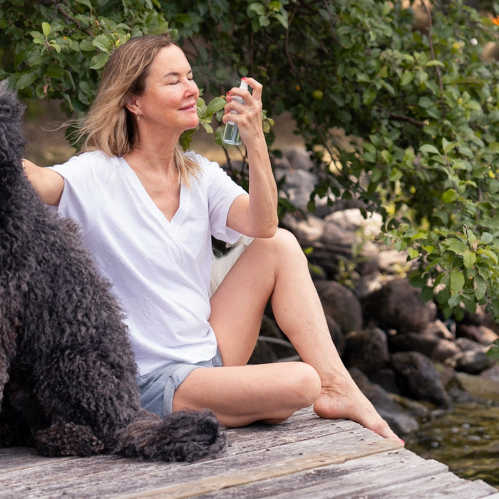 graydon moffat sits on the dock by the lake with a large curly black dog beside her. she is misting face food facial mist on her face, filled with protective and hydrating minerals to quench and soothe the skin