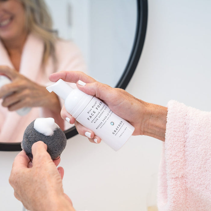 woman in fluffy robe using foaming facial cleanser, face foam, and a gently exfoliating bamboo charcoal sponge to cleanse her face